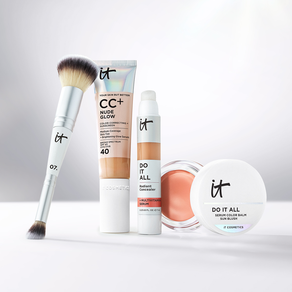 IT Cosmetics -meikkituotteita vaalealla taustalla, mukana meikkisivellin, CC+ Nude Glow, Do It All Radiant Concealer ja Do It All Serum Color Balm Sun Blush.