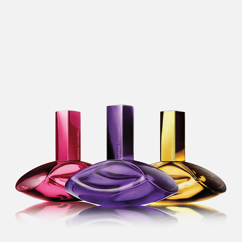 Tre färgade parfymflaskor från Calvin Klein Euphoria står sida vid sida på ljus bakgrund i rosa, lila och guld med texten "euphoria Calvin Klein" på korkarna.