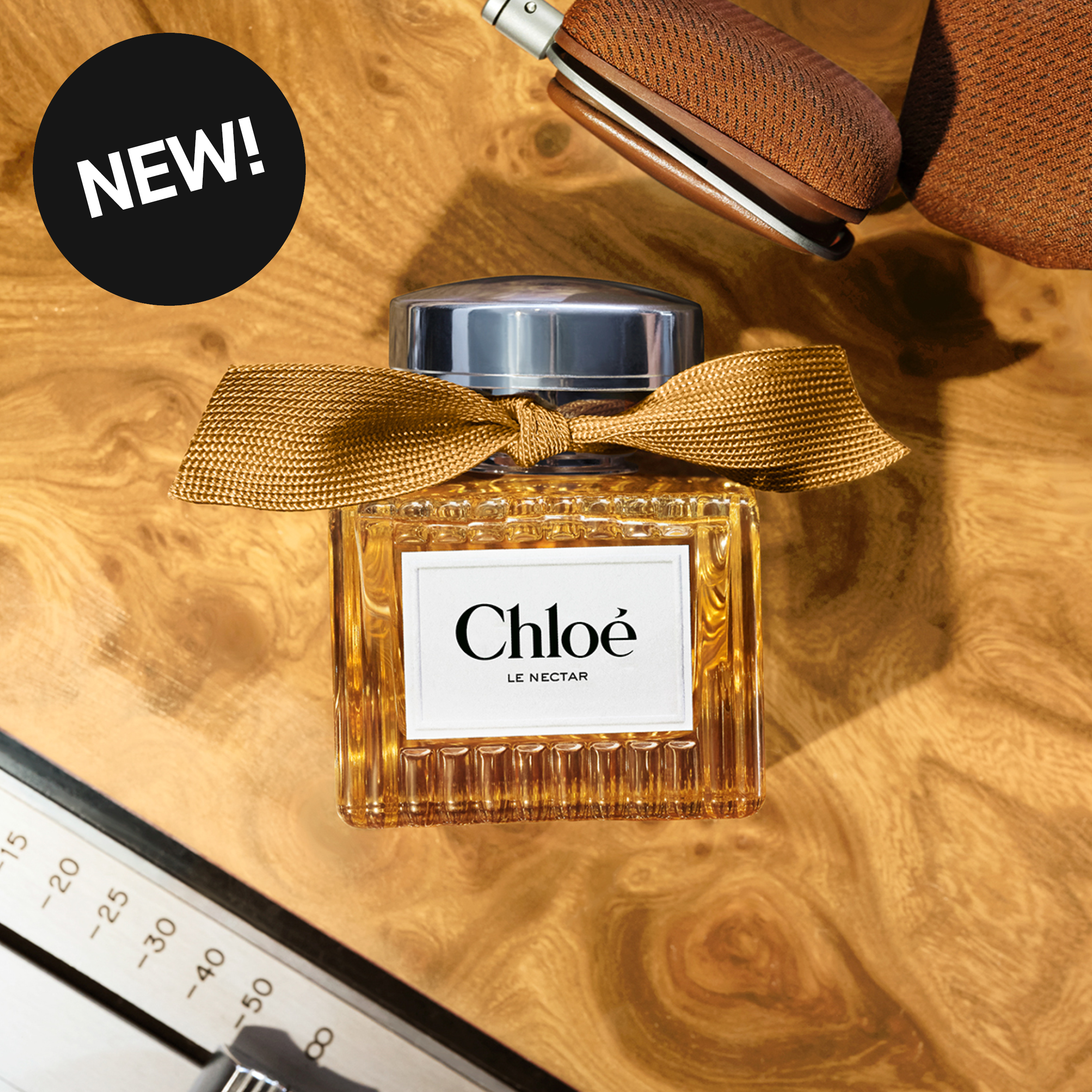 Chloé Le Nectar-parfyme i ravfarget flaske med sølvfarget kork og gullfarget sløyfe på et trebord. På flasken står «Chloé» og «LE NECTAR», og en svart sirkel viser teksten «NEW!».