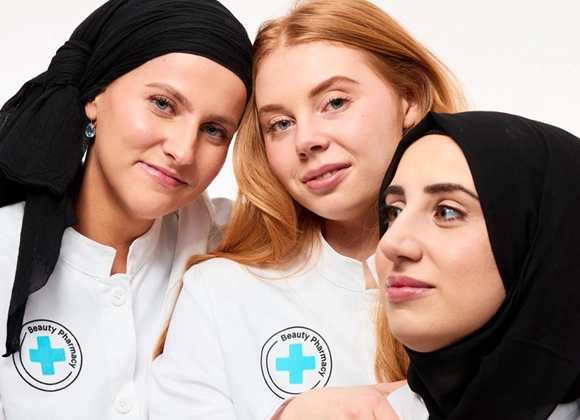 Tre kvinnor i vita arbetskläder från "Beauty Pharmacy" står nära varandra och ler mot kameran, två bär svart sjal.