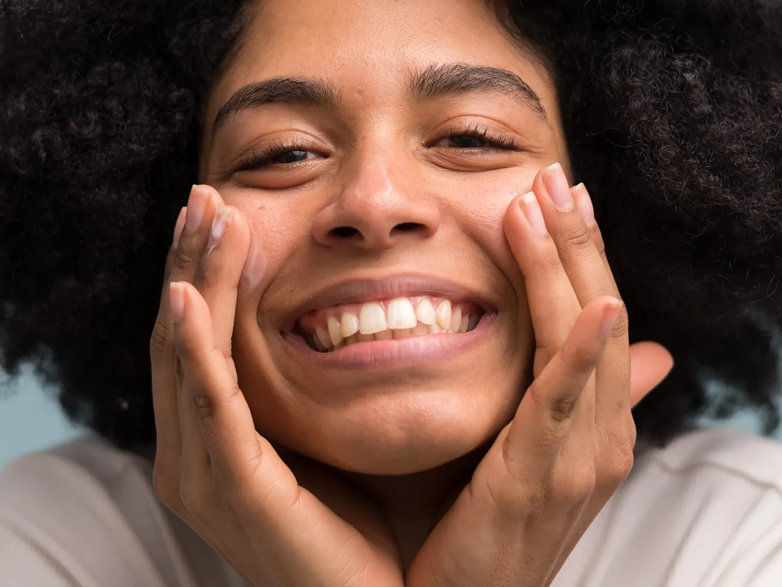 Leende person med lockigt hår som smörjer in ansiktet med hudkräm.
