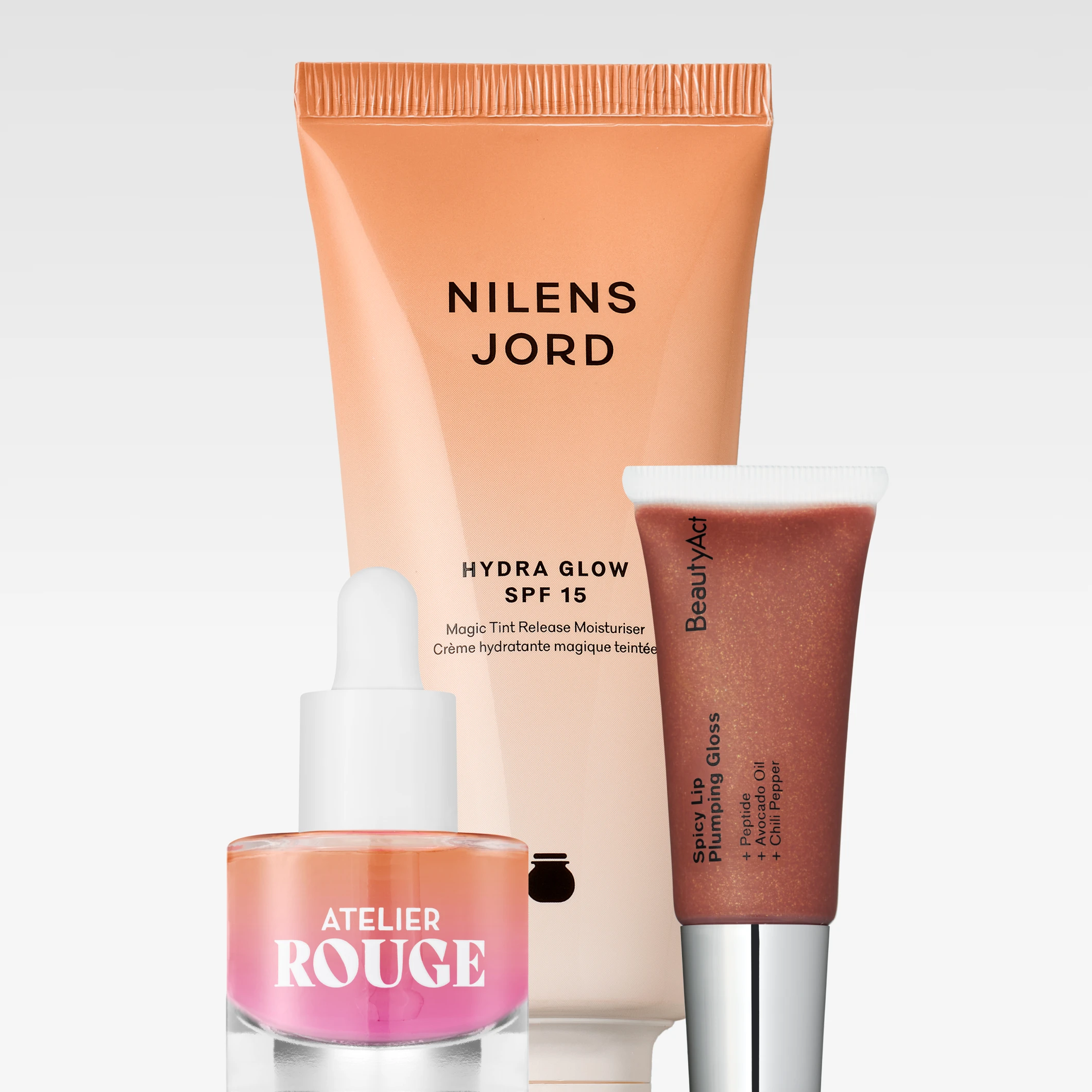 Nilens Jord Hydra Glow SPF 15 Magic Tint Release Moisturiser tillsammans med Atelier Rouge-serum och BeautyAct Spicy Lip Plumping Gloss mot ljusgrå bakgrund. På produkterna syns texterna ”NILENS JORD”, ”HYDRA GLOW SPF 15”, ”Magic Tint Release Moisturiser”, ”ATELIER ROUGE”, ”BeautyAct” och ”Spicy Lip Plumping Gloss”.