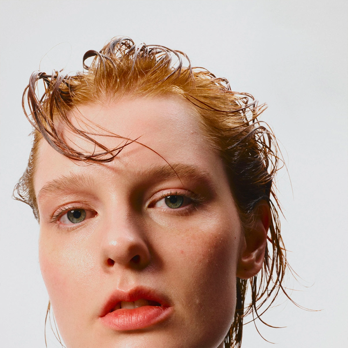 Närbild på en person med bakåtkammat våta hårstrån mot ljus bakgrund. Bilden fokuserar på hud och hårstyling med naturlig makeup och wet look-frisyr.