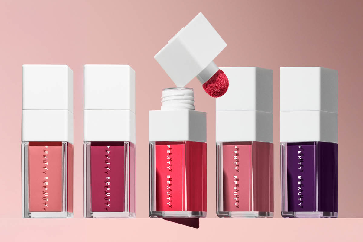 Fem flytande läpprodukter från Fenty Beauty i transparenta fyrkantiga flaskor med vita lock, i nyanser av rosa, rött och lila. En applikator med röd läppfärg hålls ovanför en öppen flaska och texten “FENTY BEAUTY” syns på förpackningarna.
