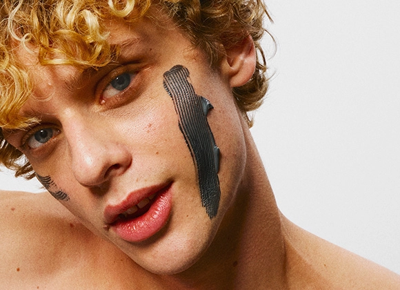 Närbild på person med lockigt blont hår och svart ansiktsmask applicerad i breda drag på kinderna.