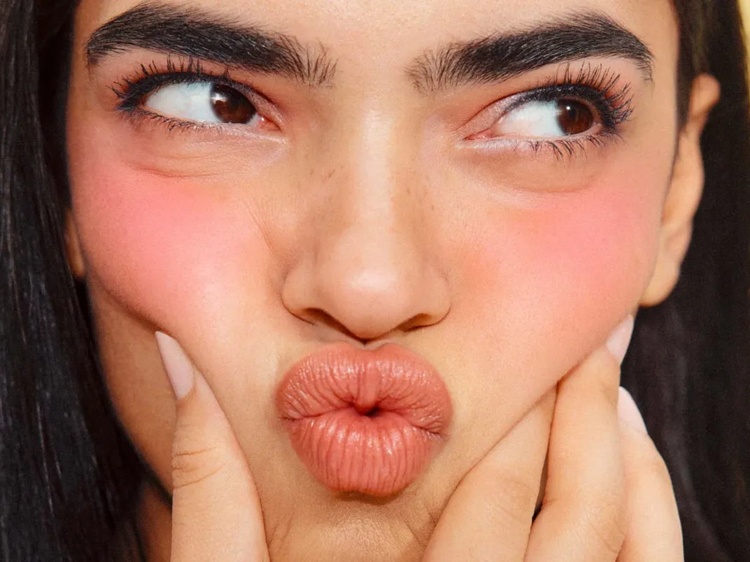 Nainen tekee pusu ilmeen samalla, kun hän puristaa punoittavia poskia käsillään.