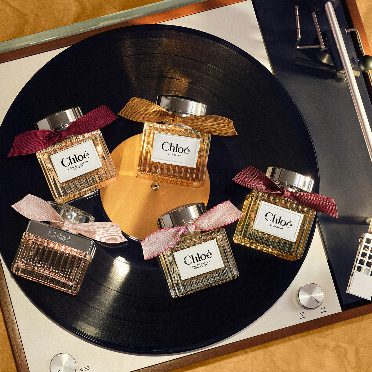 Fem Chloé-parfymer i rektangulära glasflaskor med rosetter i rosa, vinrött och guld, placerade på en svart vinylskiva på en skivspelare. På flaskorna syns texterna ”Chloé”, ”EAU DE PARFUM LUMINEUSE”, ”LE NECTAR”, ”LE PARFUM” och ”L’EAU DE PARFUM INTENSE”.