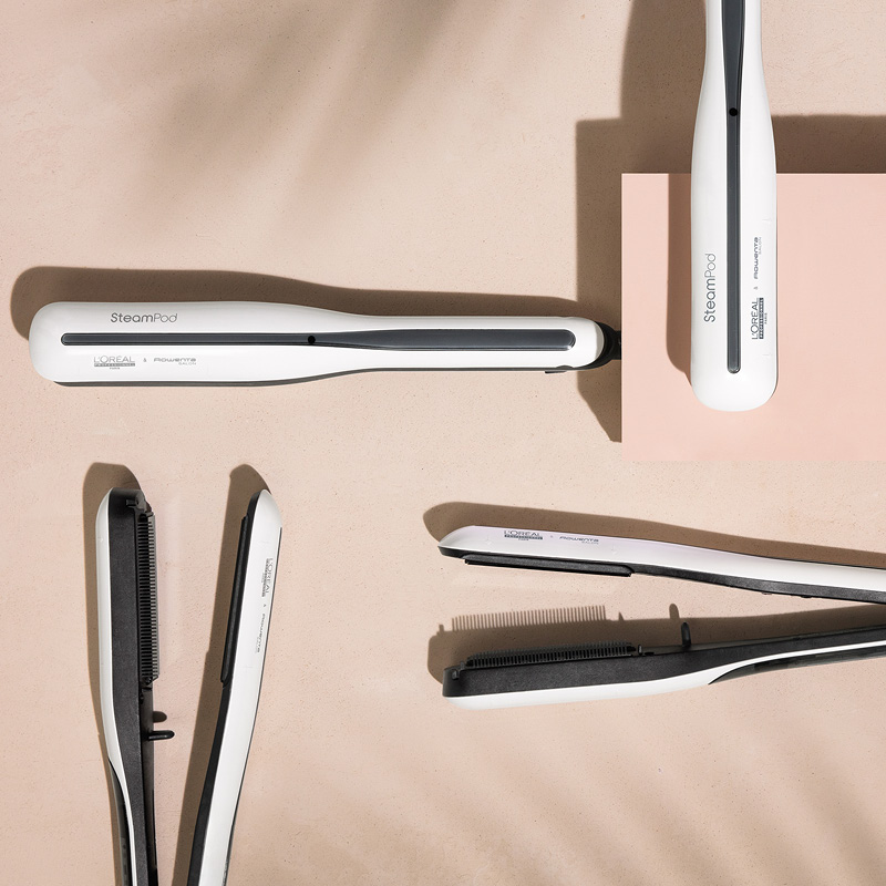 Fem hvite L'Oréal Professionnel SteamPod rettetang er spredt ut på en beige flate. Noen av rettetangene er åpne, mens andre er lukket. Bakgrunnen har en skyggelagt effekt.