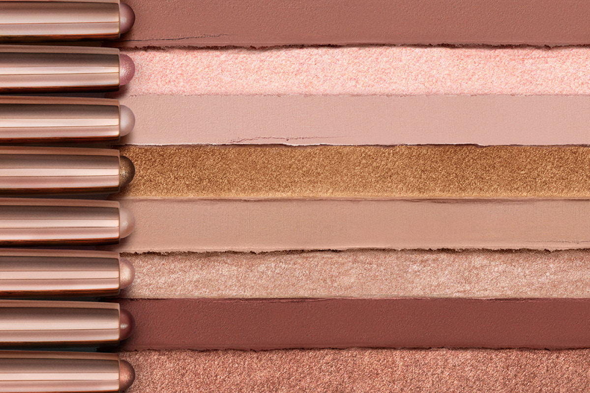 Närbild på ögonskuggspennor i nude-, beige-, brun- och champagnefärgade nyanser med både matta och skimrande finishes. Makeupprodukterna är applicerade i horisontella drag mot en neutral bakgrund och visar olika toner för ögonmakeup.