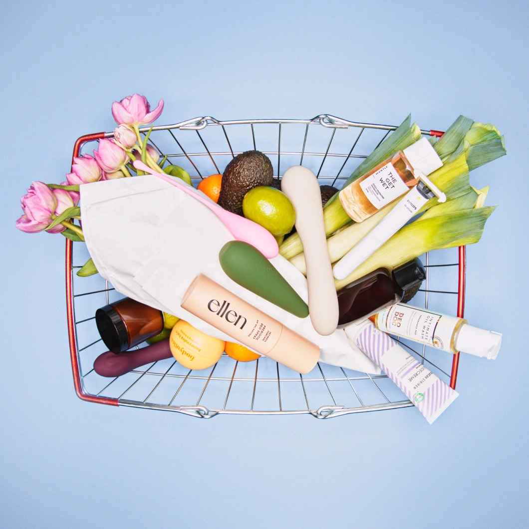 En handlevogn fylt med wellness- og intimpleieprodukter vises mot lys blå bakgrunn. Handlevognen inneholder blant annet blomster, avokado, sitrusfrukter, sexleketøy og produkter fra ellen, DeoDoc og The Get Wet.