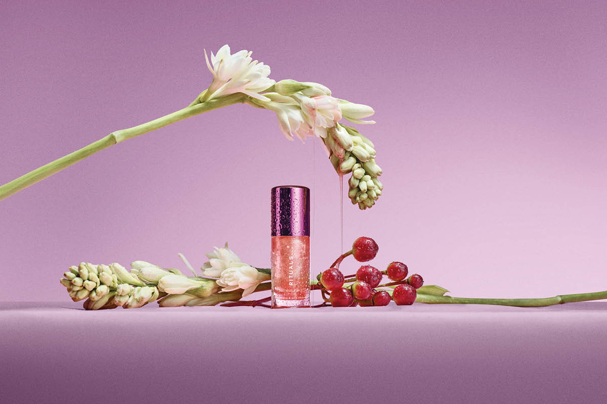 Rituals leppeprodukt i en liten flaske med skimrende rosa innhold og teksten “RITUALS”, plassert mellom hvite blomster og røde bær mot rosa bakgrunn.