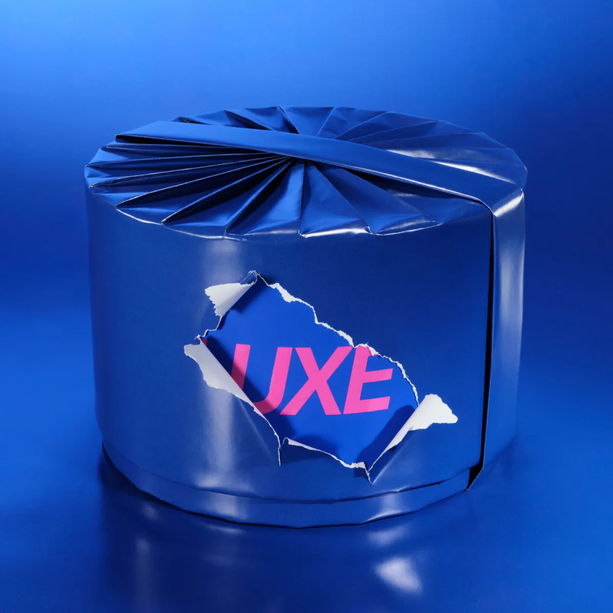 Siniseen kiiltävään paperiin kääritty pyöreä pakkaus sinisellä taustalla. Repeytyneestä kohdasta näkyy pinkki ”LUXE”-teksti.