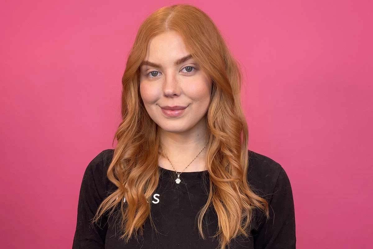Henkilö, jolla on pitkä, punainen tukka, seisoo vaaleanpunaisen taustan edessä ja on pukeutunut mustaan paitaan, jossa lukee "KICKS". Hiukset on muotoiltu löysiksi kiharoiksi.