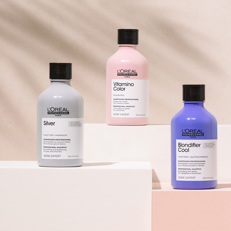 Tre sjampoer fra L'Oréal Professionnels Serie Expert er stilt opp på hvite kuber mot en beige bakgrunn. En flaske er grå, en er rosa og en er lilla. Alle flaskene har svart lokk og hvite etiketter med svart tekst.