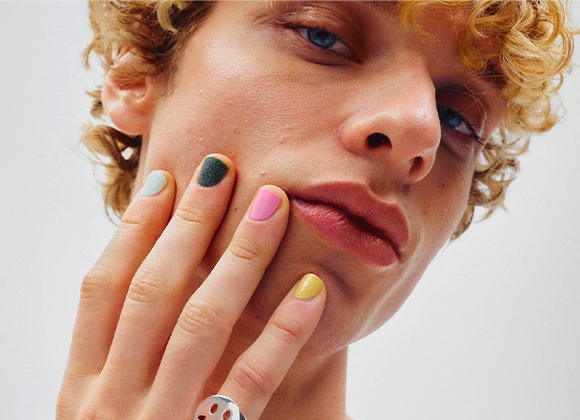 Lähikuva kasvoista, käsi poskella ja kynnet lakattu eri väreillä kuten pinkki, keltainen, vihreä ja vaaleansininen. Lyhyet kiharat hiukset, luonnollinen ihon tekstuuri ja neutraali tausta.
