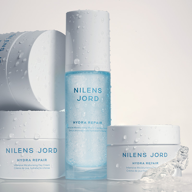 Nilens Jord Hydra Repair-produkter som inkluderer intensivt fuktighetsgivende dagkrem og serum med mikro-kapsler. Produktene er i hvite og blå emballasjer, og de er dekket med vanndråper.