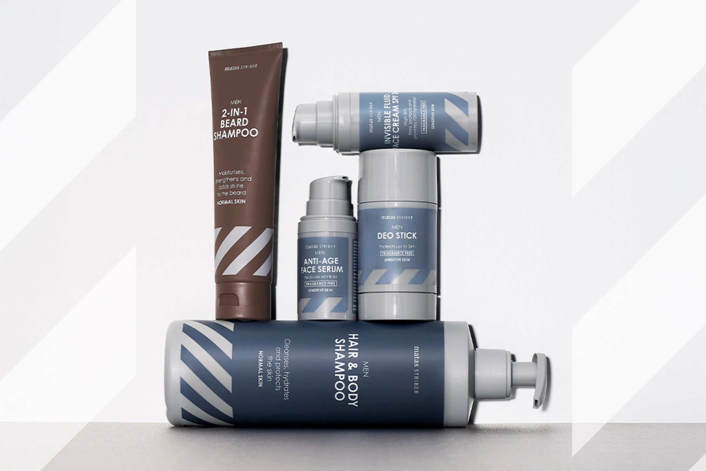 Matas Striber MEN -tuotteita aseteltuna vaaleanharmaata taustaa vasten, jossa on hillittyjä vinoraitoja. Tuotteissa näkyvät tekstit sisältävät "2-IN-1 BEARD SHAMPOO", "ANTI-AGE FACE SERUM", "DEO STICK", "INVISIBLE FLUID FACE CREAM SPF" ja "HAIR & BODY SHAMPOO".