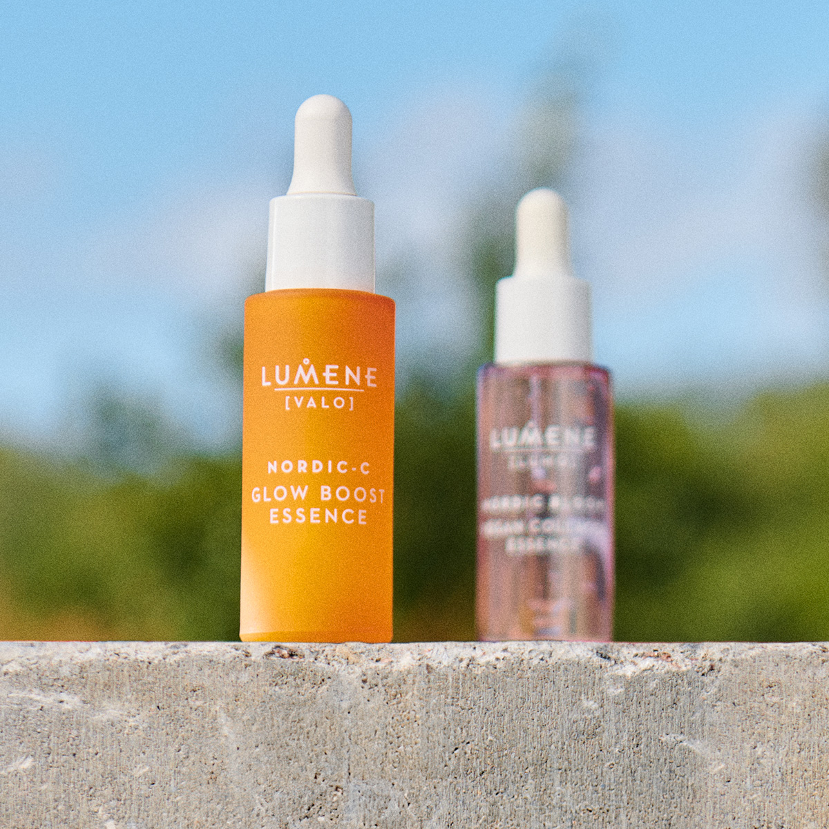 To flasker med Lumene hudpleieprodukter står på en betongoverflate utendørs. Den fremste flasken er oransje og inneholder Nordic-C Glow Boost Essence, mens den bakre flasken er gjennomsiktig med rosa væske.