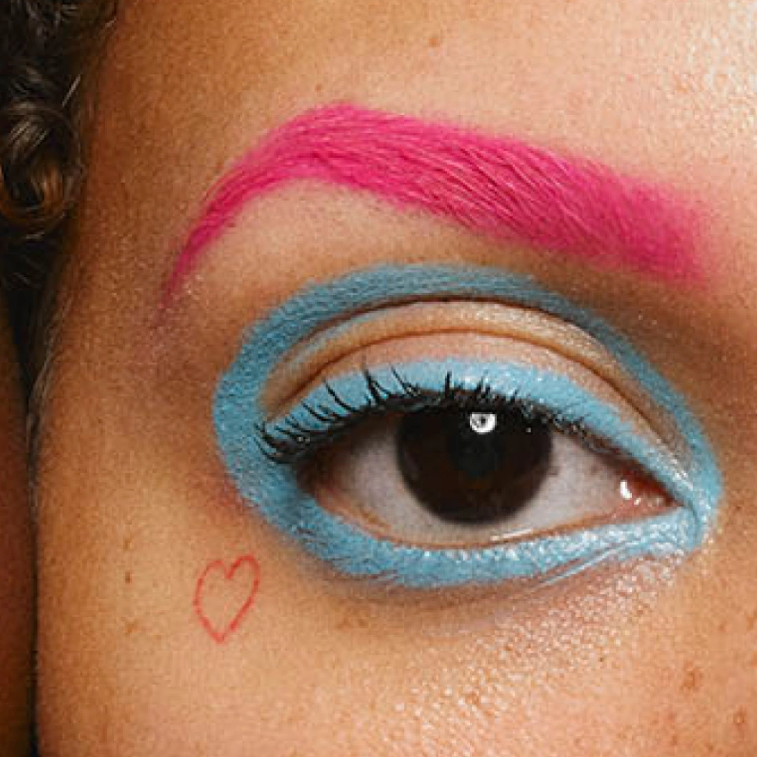 Närbild på ett öga med intensivt turkosblå ögonmakeup runt ögat, matchande blå ögonskugga och en starkt rosa målad ögonbrynsliknande detalj ovanför. Ett litet rött hjärta är ritat under ögat.