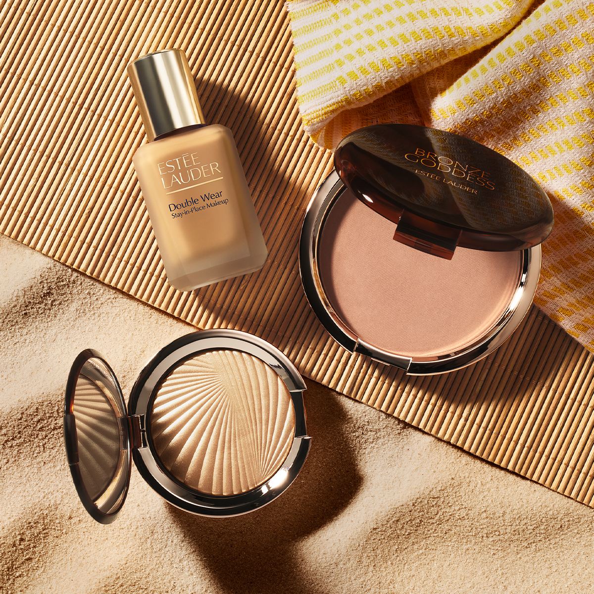 Estée Lauder-sminkeprodukter er plassert på sand ved siden av en bambusmatte og et stripete håndkle. Bildet viser foundationen «ESTÉE LAUDER Double Wear Stay-in-Place Makeup», et åpent pudder med teksten «BRONZE GODDESS ESTÉE LAUDER» samt en åpen highlighter i gulltone.