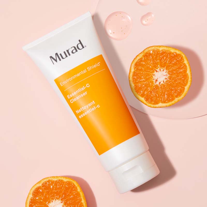 Rengöring från Murad placerad på en rosa bakgrund. Tuben är vit med en orange etikett som visar produktnamnet "Environmental Shield Essential-C Cleanser". Runt tuben ligger två halvor av en apelsin och några droppar av produkten, vilket framhäver dess C-vitamin innehåll och fräscha känsla.