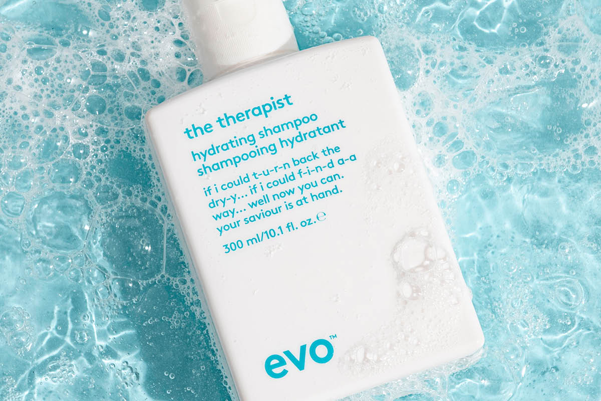 Lähikuva EVO “the therapist” hydrating shampoo -tuotteesta valkoisessa pullossa sinisen veden, vaahdon ja kuplien päällä. Pakkauksessa näkyvät selkeästi tekstit “the therapist”, “hydrating shampoo”, “shampooing hydratant”, “300 ml/10.1 fl. oz.” ja “evo”.