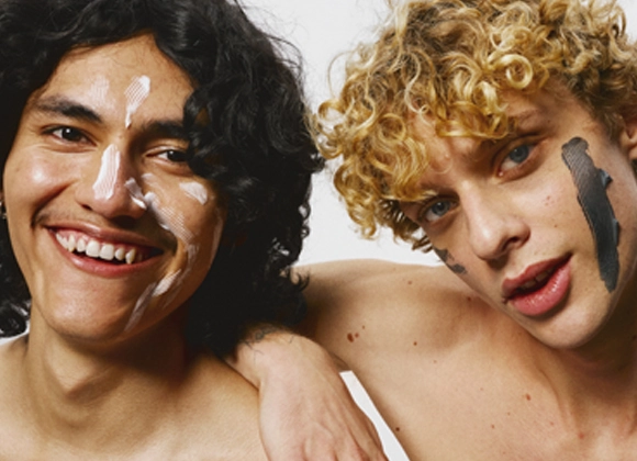 Två leende unga män med hudvårdsprodukter applicerade i ansiktet poserar tillsammans mot vit bakgrund.