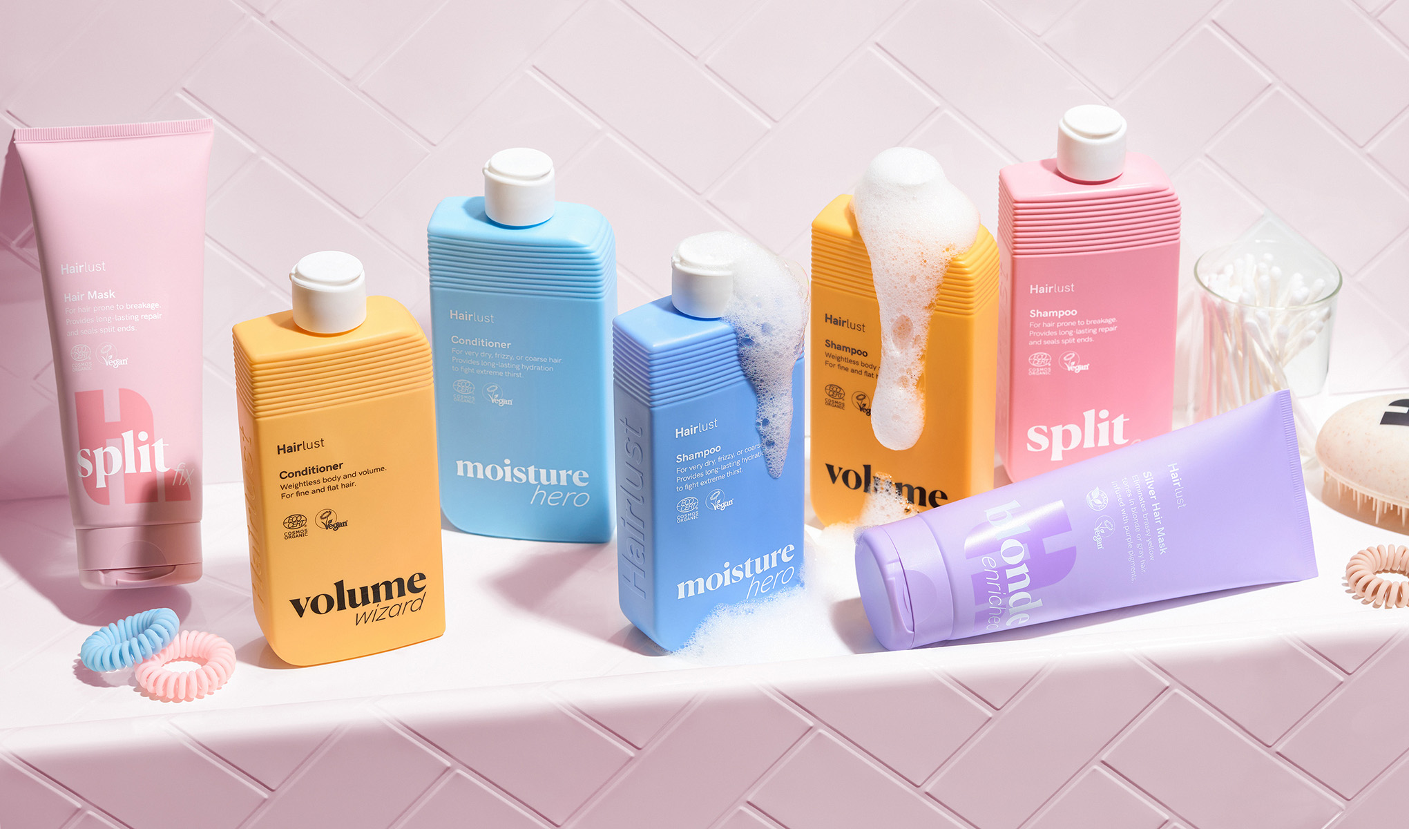 En samling hårpleieprodukter fra Hairlust er stilfullt arrangert på en hvit flate foran en rosa flislagt vegg. Produktene er i fargerike forpakninger, som rosa, blå, gul og lilla. Noen av flaskene har skum på seg, noe som gir en følelse av bruk. Rundt produktene er det hårbånd og en børste. Designet er lekent og fargerikt, med fokus på å fremheve produktenes ulike funksjoner og fordeler.