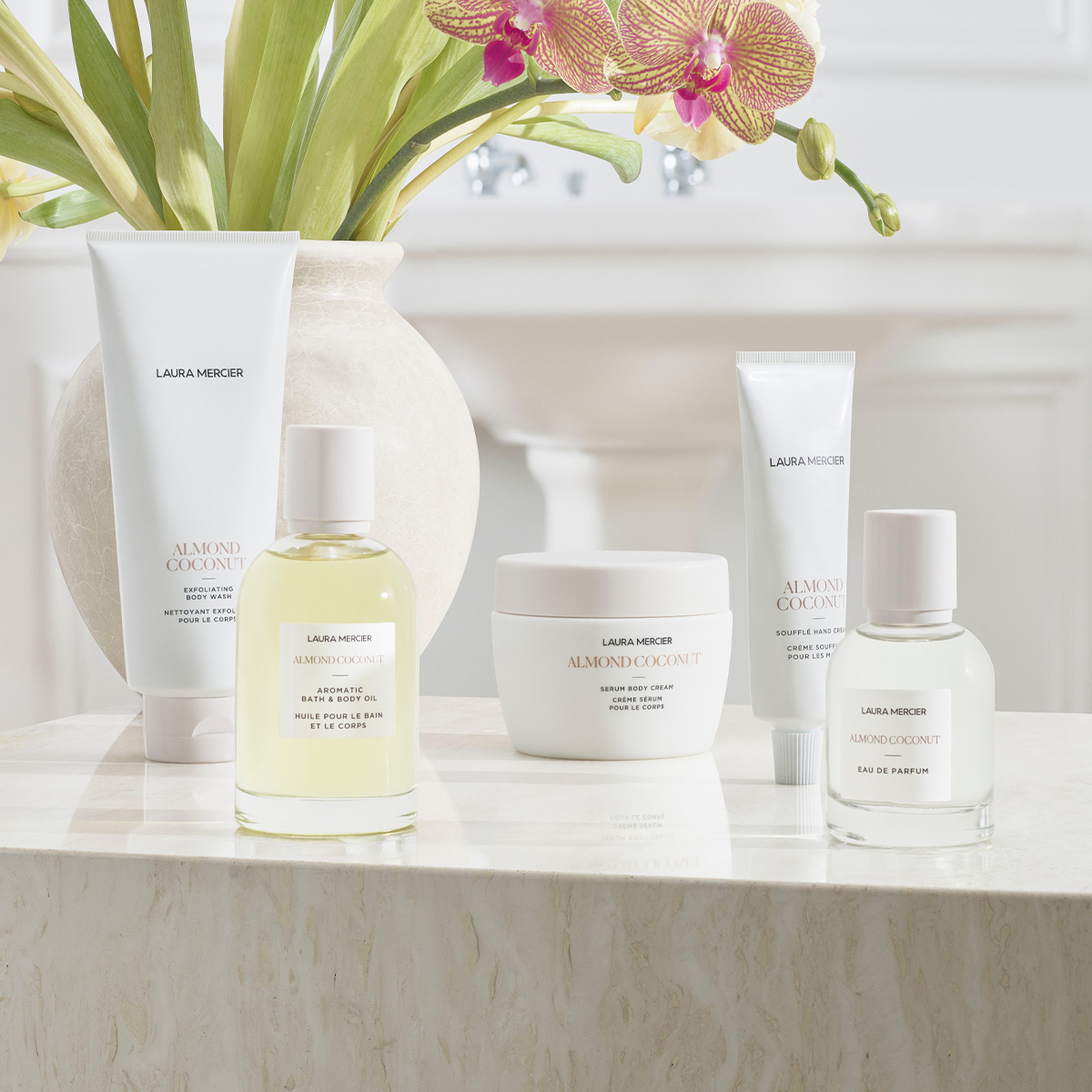 Laura Merciers Almond Coconut-produkter för kroppen, placerade på en marmoryta med en vas med blommor i bakgrunden.