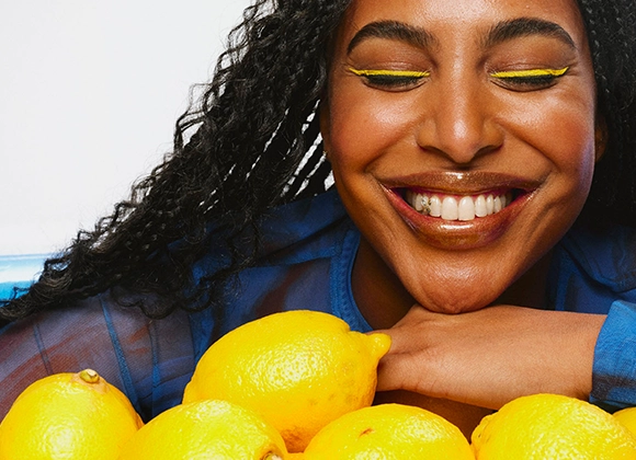 Person med gul eyeliner, glansigt läppglans och blå topp lutar sig över en rad gula citroner, i en färgstark beautybild med fokus på makeup som eyeliner och lipgloss.