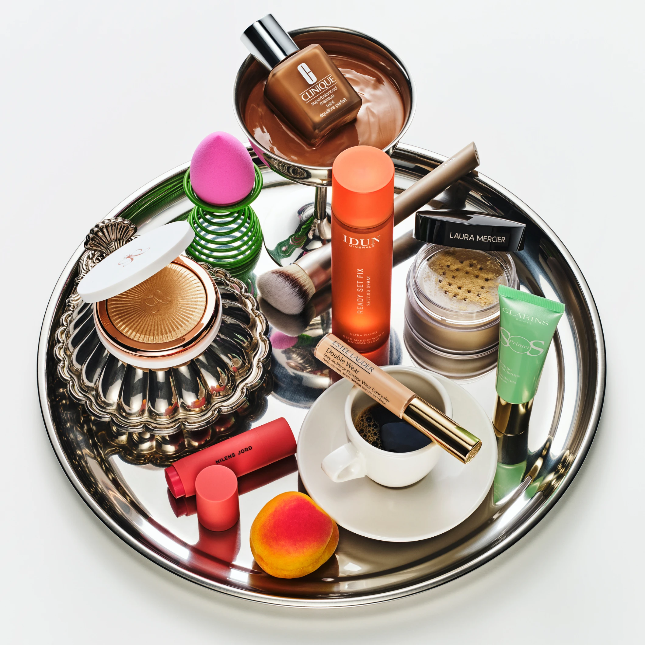 en silverfärgad bricka med flera sminkprodukter, bland annat foundation i flaska, löspuder i burk, settingspray, primer, sminkborste, makeupsvamp, läpprodukter och en kaffekopp.
