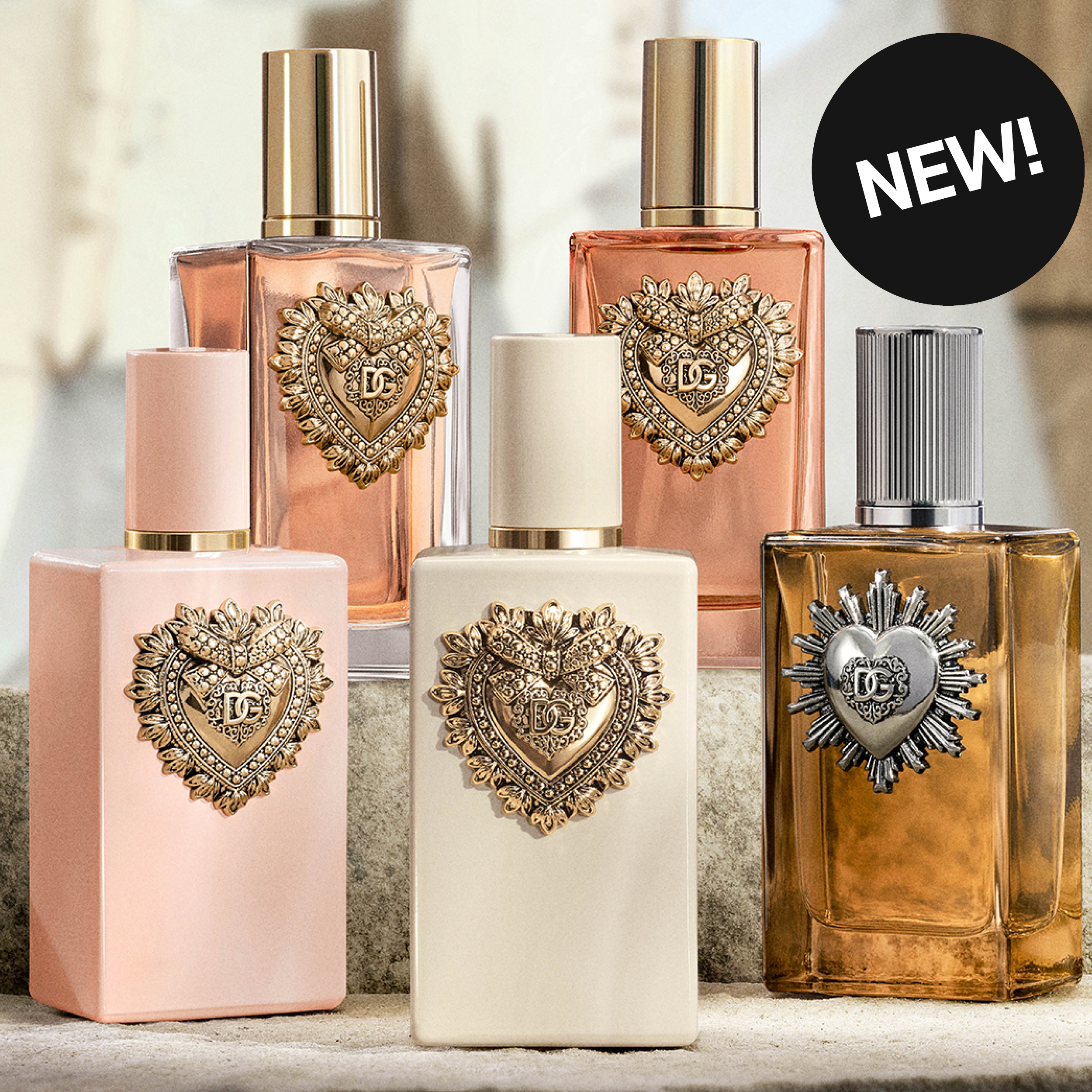 Fem Dolce & Gabbana-parfymeflasker i rosa, kremfarget og ravfarget glass med dekorative hjerteformede DG-emblemer i gull og sølv. En svart sirkel viser teksten «NEW!».