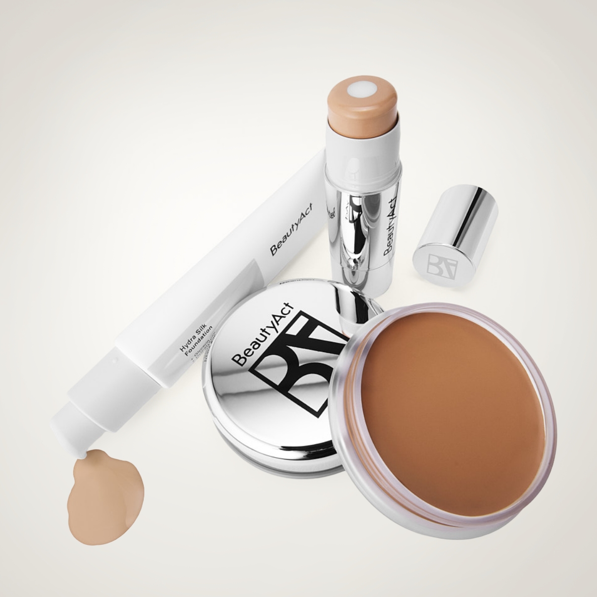 BeautyAct makeup-produkter med foundation og base: tube merket “BeautyAct Hydra Silk Foundation”, stickfoundation, kompakt med BA-logo og krukke med kremet foundation i brun nyanse mot lys bakgrunn.