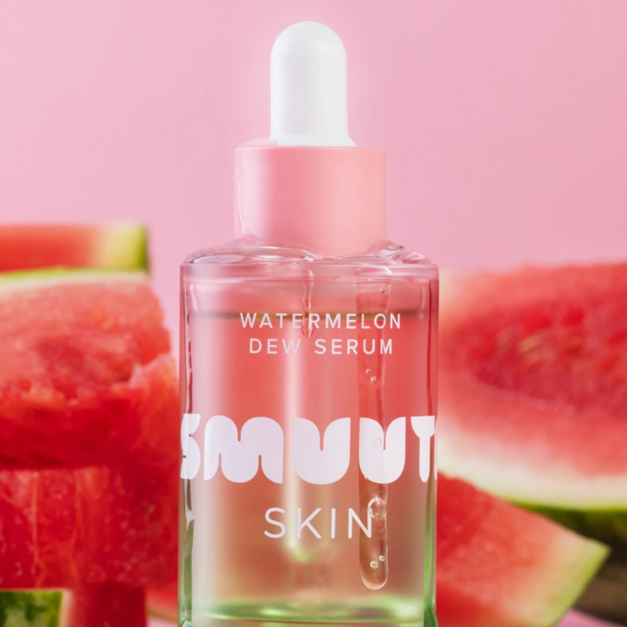 Smuuti Skin Watermelon Dew Serum i transparent flaske med rosa pipette mot rosa bakgrunn, omgitt av vannmelonskiver. Synlig tekst på flasken: «WATERMELON DEW SERUM», «SMUUTI» og «SKIN».