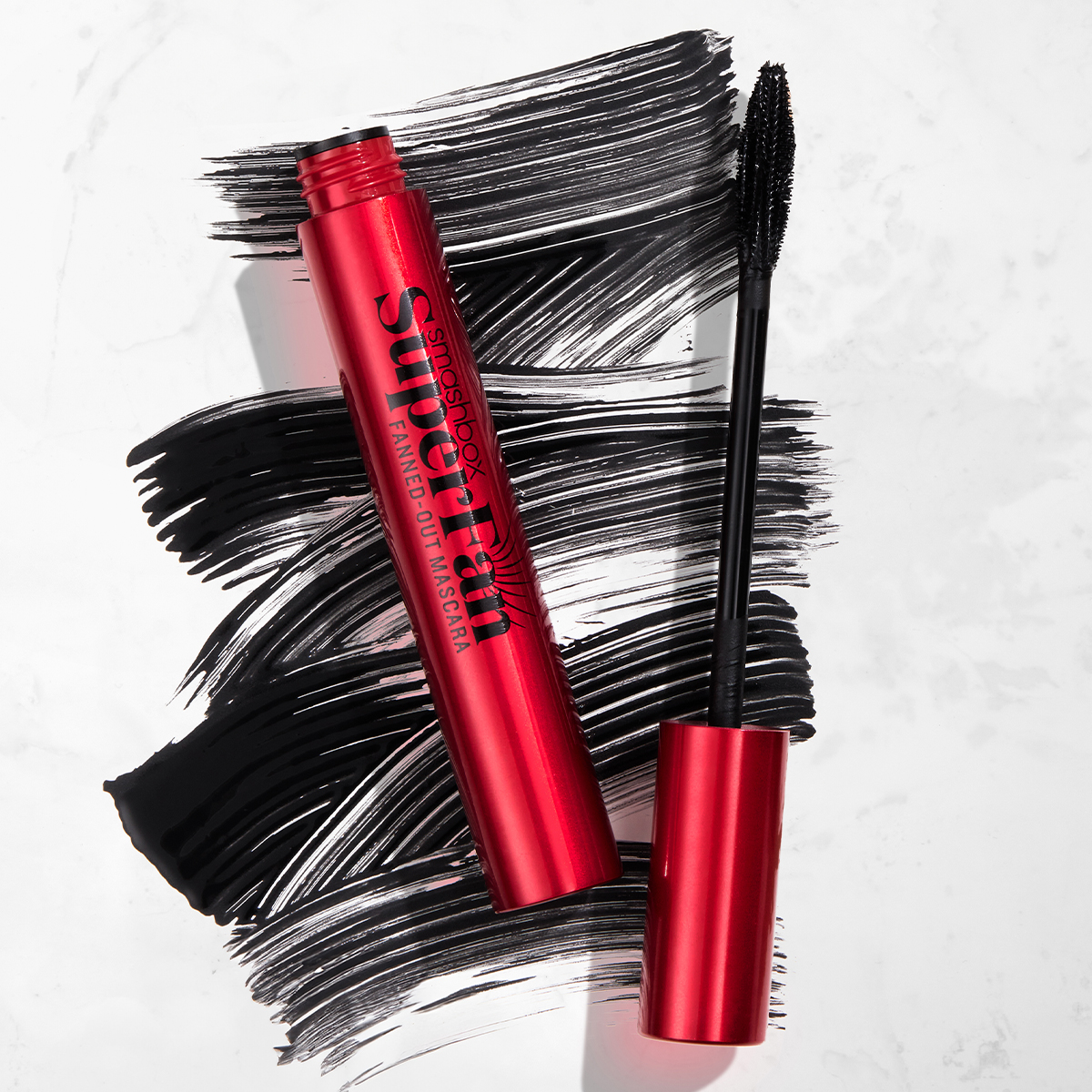 Smashbox Superfan Fanned-Out Mascara i en rød tube, med mascarabørsten ved siden av og svarte mascarastrøk i bakgrunnen.