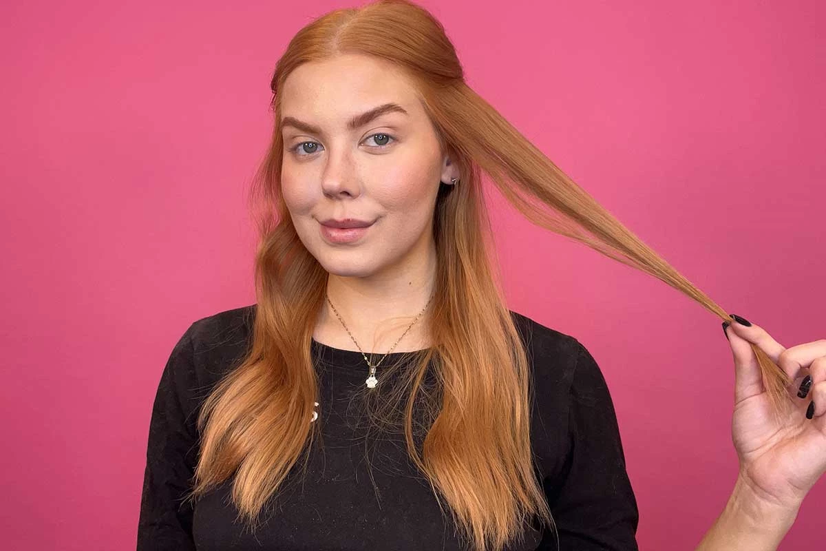 Henkilö, jolla on pitkä, punainen tukka, seisoo vaaleanpunaisen taustan edessä ja on pukeutunut mustaan paitaan, jossa lukee "KICKS". Henkilö pitää hiussuortuvaa kädessään.