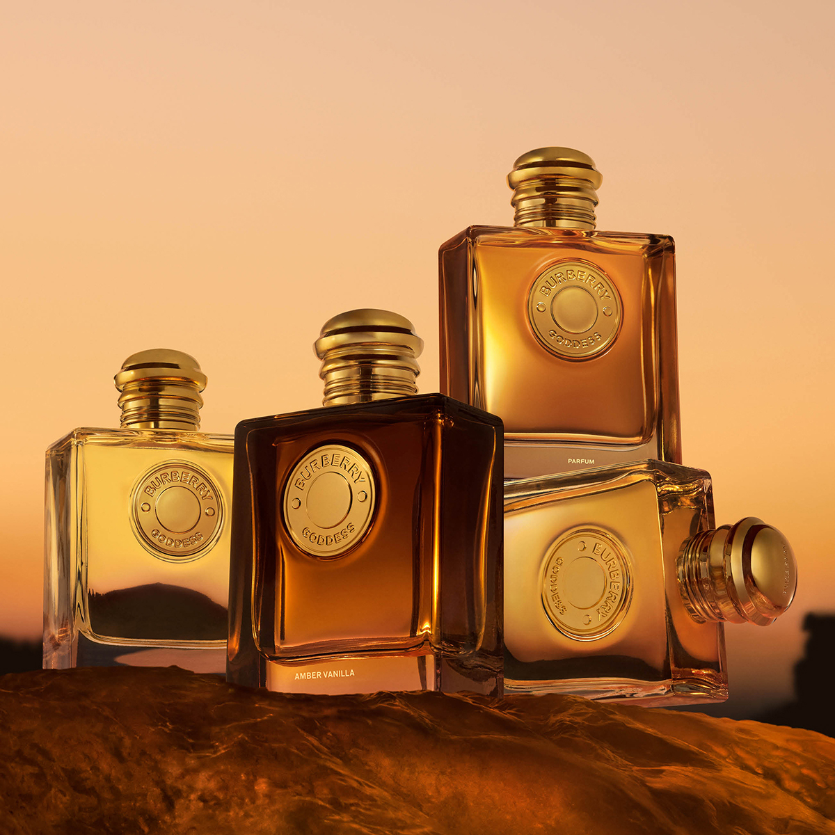 Fire Burberry Goddess-parfymer i rektangulære glassflasker med gullfargede detaljer, arrangert mot en varm oransje bakgrunn. Teksten «BURBERRY GODDESS», «PARFUM» og «AMBER VANILLA» vises på flaskene.