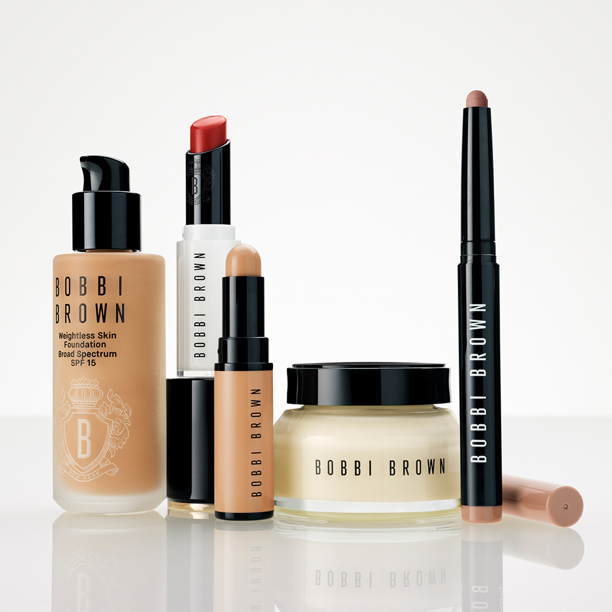 Useita Bobbi Brown -meikkituotteita on esillä vaaleaa taustaa vasten: Weightless Skin Foundation Broad Spectrum SPF 15, huulipuna, meikkipuikko, kasvovoide purkissa ja meikkikynä. Näkyvä teksti: "BOBBI BROWN", "Weightless Skin Foundation", "Broad Spectrum SPF 15", "SPF 15" ja "NEW YORK".