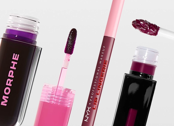 Nærbilde av flere flytende leppeprodukter mot lys bakgrunn. Åpne beholdere med applikatorer er synlige, fylt med pigment i rosa og mørkelilla nyanser. En applikator med produkt er plassert over en av beholderne.