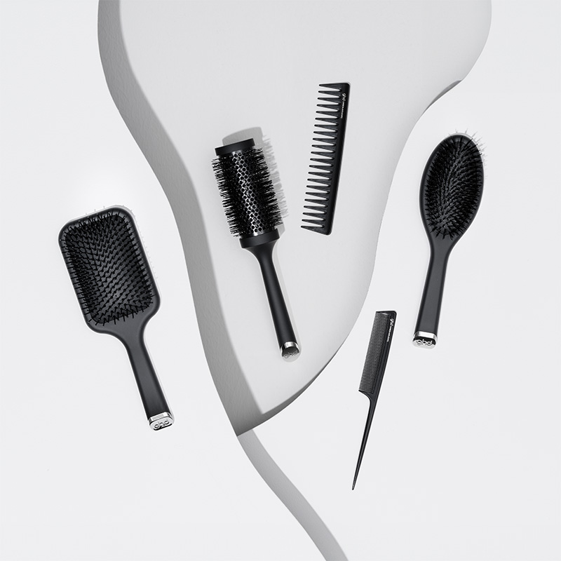 En samling av svarte GHD-hårbørster og kammer er stilfullt arrangert på en hvit bakgrunn med en bølgende form. Designet er elegant og moderne, med fokus på detaljer og funksjonalitet.