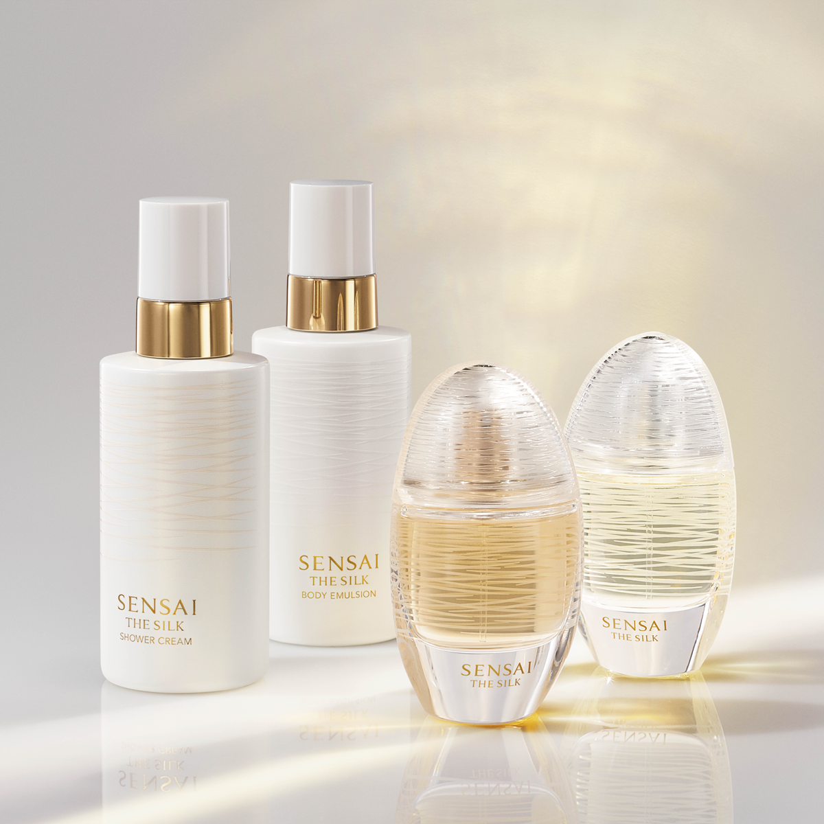 Fire produkter fra SENSAI The Silk vises mot en lys bakgrunn: Shower Cream, Body Emulsion og to parfymeflasker fra The Silk-serien. Produktene har hvit og transparent design med gullfargede detaljer som gir et eksklusivt uttrykk av parfyme, body lotion og kroppspleie.