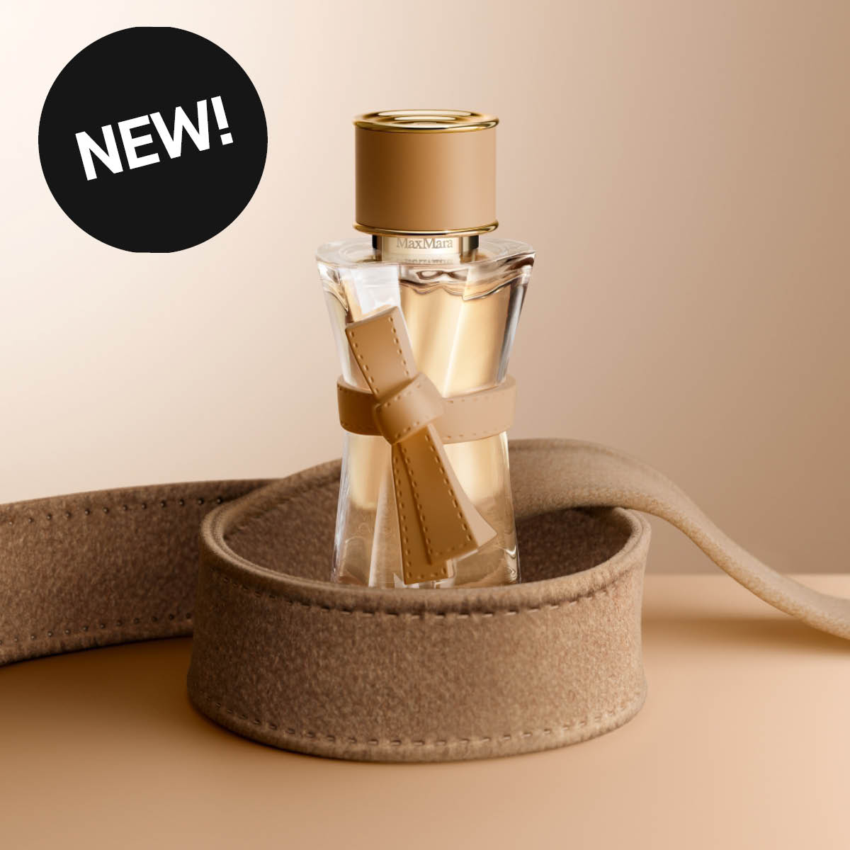Max Mara-parfyme i gjennomsiktig flaske med gullfarget kork og beige sløyfedetalj, plassert i et mykt beige etui. Svart sirkel med den hvite teksten «NEW!».