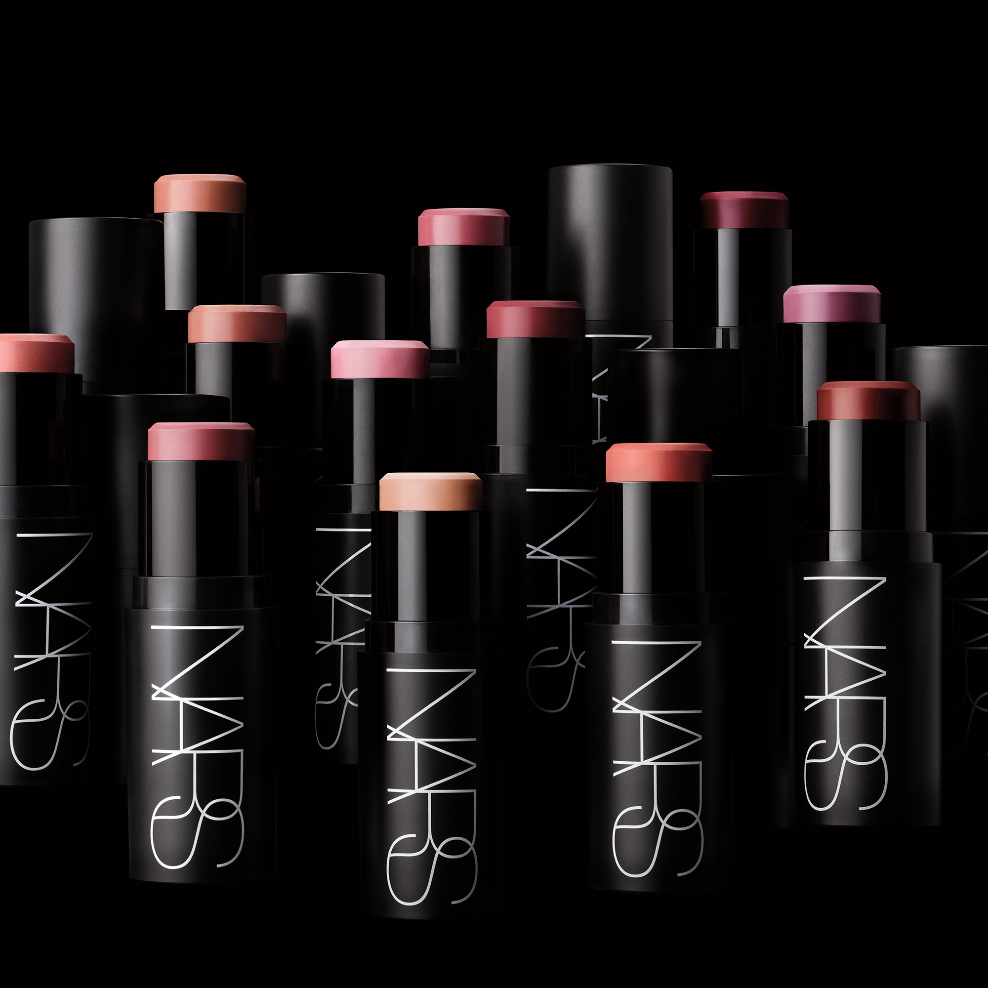 Useita NARS cream blush -puikkoja vaaleanpunaisen, persikan, nude-sävyjen ja marjasävyjen väreissä on aseteltu mustaa taustaa vasten. Mustissa pakkauksissa näkyy valkoinen “NARS”-logo, ja esillä on useita voidemaisia poskipunasävyjä.