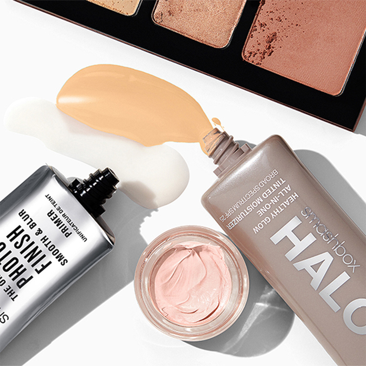 En samling Smashbox-sminkeprodukter plassert på en hvit flate. En åpen palett er synlig i øvre del av bildet. Ved siden av en sølvfarget tube og en beige tube står en krem som har rent ut. I midten står en liten krukke med rosa krem. Produktene har sølvfarget, beige og rosa emballasje.