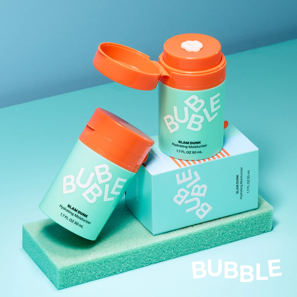Två Bubble Slam Dunk återfuktande krämer i turkos förpackning med orange lock, varav en är öppen med kräm synlig. Produkterna är stylade på en färgad yta mot en ljusblå bakgrund.