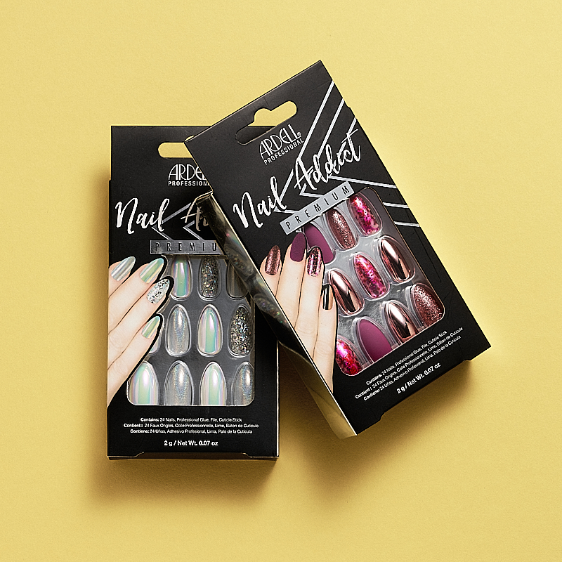 Förpackningar med Ardell Professional Nail Addict Premium lösnaglar. Den ena förpackningen innehåller naglar i holografiska och glittriga silverfärger, medan den andra innehåller naglar i rosa, lila och glittriga färger. Förpackningarna är placerade på en gul bakgrund.