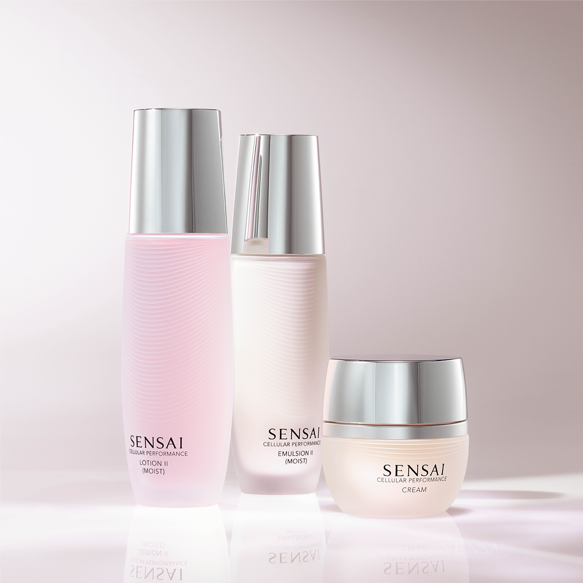 Tre hudpleieprodukter fra SENSAI Cellular Performance står foran en lys rosa bakgrunn: Lotion II (Moist), Emulsion II (Moist) og Cream. Produktene har myke rosa og hvite toner med sølvfargede lokk og gir et eksklusivt uttrykk for fuktighetsgivende skincare og ansiktskrem.