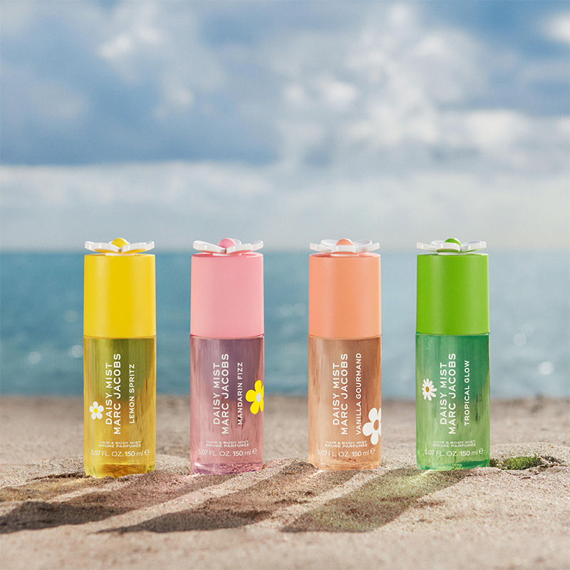 Fyra Marc Jacobs Daisy Mist Hair & Body Mist står på sand framför havet. Flaskorna är Lemon Spritz, Mandarin Fizz, Vanilla Gourmand och Tropical Glow i gula, rosa, persikofärgade och gröna nyanser med blomformade korkar och synlig produkttext.