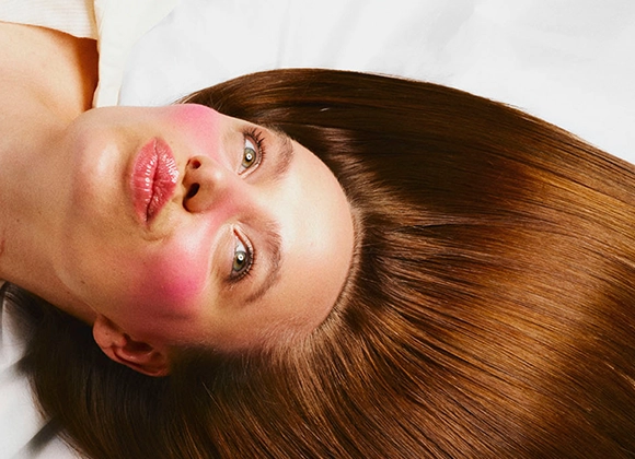 Närbild av en person som ligger ner med huvudet vänt åt sidan. Personen har långt, rakt brunt hår som breder ut sig runt huvudet. Ansiktet är synligt med rosatonad makeup på kinder och näsa samt glansiga läppar. Ljusa ögon och vit bakgrund.