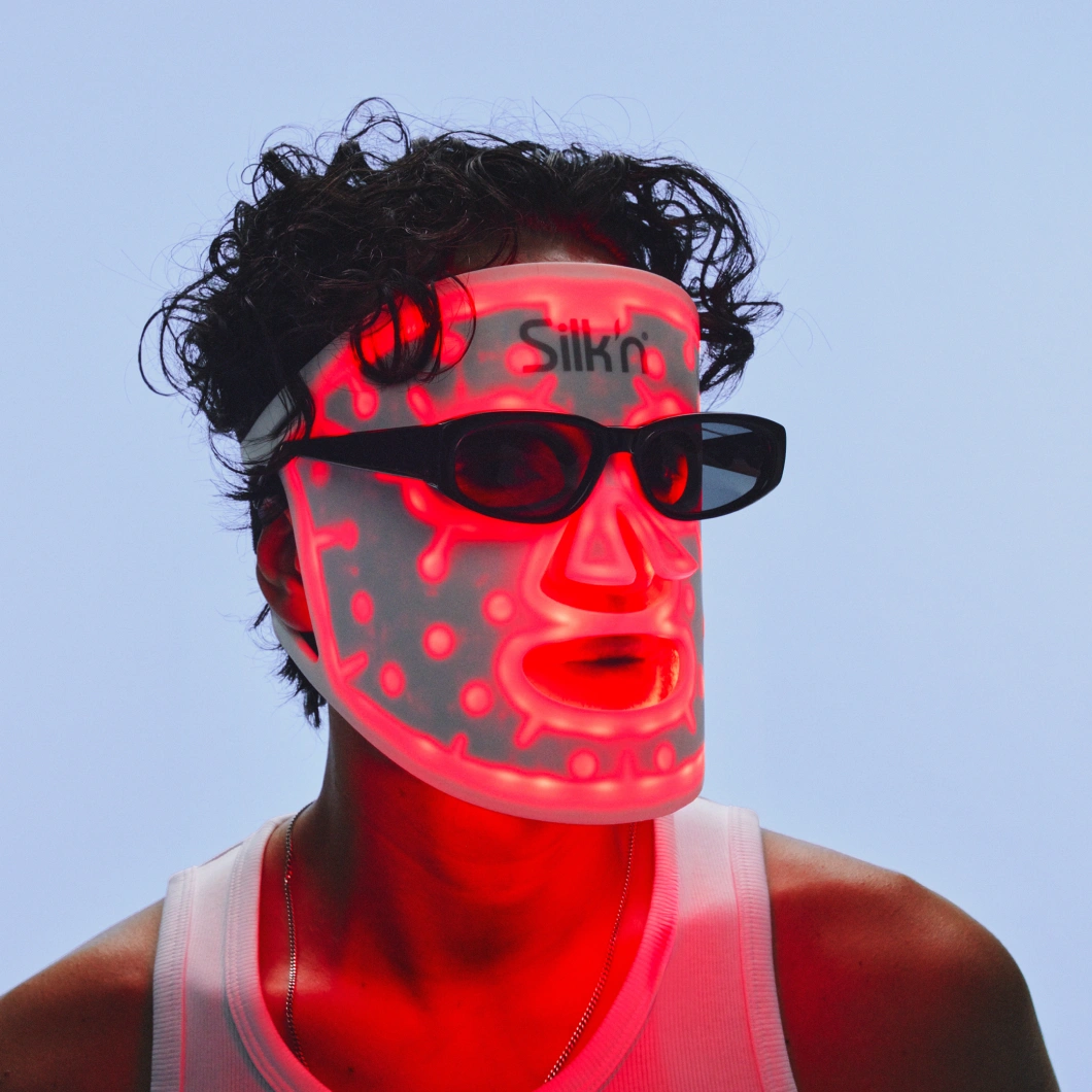En person med mørkt krøllete hår bruker en opplyst Silkn LED-maske og svarte solbriller mot lys blå bakgrunn. Det røde LED-lyset lyser gjennom masken og ansiktet.